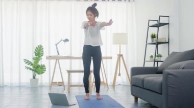 Spor giyinen genç Koreli bayan evde yoga videosu izlemek için spor yapıyor ve dizüstü bilgisayar kullanıyor. Kişisel antrenörle uzak eğitim, sosyal mesafe, çevrimiçi eğitim kavramı.