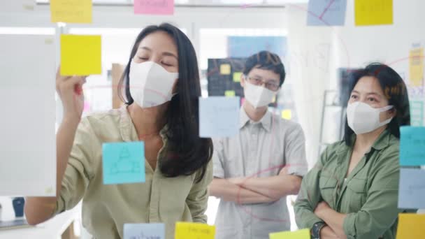 アジアの若いビジネスマンたちは オフィスで仕事をしているときに データを共有し ガラスの壁を医療用フェイスマスクで書き戻すビジネスブレインストーミング会議について話し合っています コロナウイルスの後の生活と仕事 — ストック動画