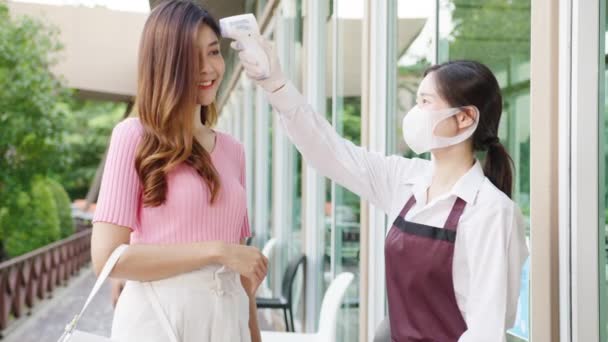 在进入之前 年轻的亚洲女餐馆工作人员戴着防护面罩 在顾客的额头上使用红外温度计检测仪或温度枪 电晕病毒后的新生活方式 — 图库视频影像
