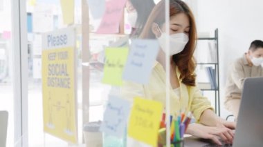 Asyalı kadınlar, dizüstü bilgisayar kullanırken ve ofiste akrilik bölünme ile ayrılırken, virüsün önlenmesi için yeni normal durumlarda sosyal mesafeler için yüz maskesi takarlar. Corona virüsünden sonra hayat ve iş.
