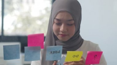 Asyalı Müslüman kadın yeni normal ofisindeki cam tahtaya yapışkan bir not yazdı. Evden çalışmak, uzaktan çalışmak, kişisel izolasyon, sosyal uzaklık, koronavirüs önleme için karantina.