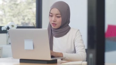 Güzel Asyalı Müslüman kadın gündelik kıyafetleri modern normal ofiste dizüstü bilgisayarla çalışıyor. Evden çalışmak, uzaktan çalışmak, kişisel izolasyon, sosyal uzaklık, koronavirüs önleme için karantina.