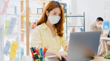 Asyalı kadınlar, dizüstü bilgisayar kullanırken ve ofiste akrilik bölünme ile ayrılırken, virüsün önlenmesi için yeni normal durumlarda sosyal mesafeler için yüz maskesi takarlar. Corona virüsünden sonra hayat ve iş.