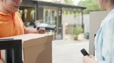 Genç posta kuryesi, paketleri müşteriye göndermek için elinde tutuyor, Asyalı kadınlar ise dışarıda paket teslim almak için cep telefonuna QR kodu ile imza atıyor ve ödeme yapıyor. 4k Ağır çekim.