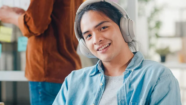 Young Asian designer man listening to music on headphones, looking at camera and smiling in modern office. Group of young college students in smart casual wear on campus. Coworker teamwork concept.