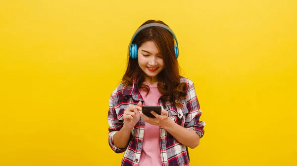Young Asian lady wearing wireless headphones listening to music from smartphone with cheerful expression in casual clothing and looking at camera over yellow background. Facial expression concept.