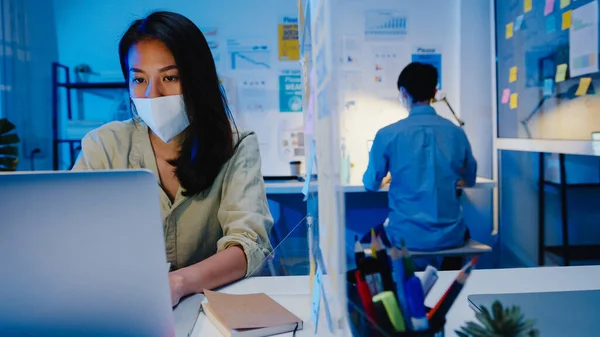 Happy Asia businesswoman wearing medical face mask for social distancing in new normal situation for virus prevention while using laptop back at work in office night. Life and work after coronavirus.