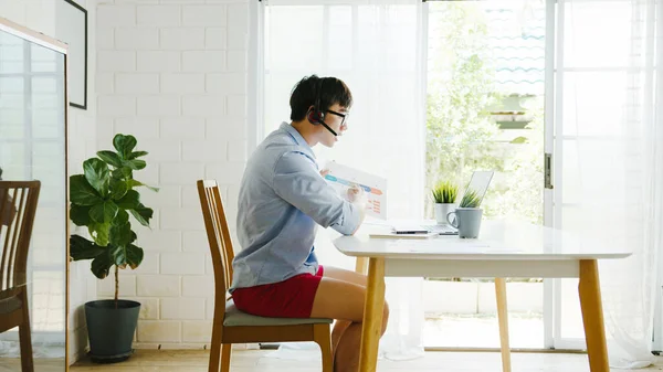 Asia businessman dressed in shirt and shorts use laptop talk to colleagues in video call while work from home at living room. Self-isolation, social distancing, quarantine for corona virus prevention.