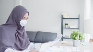 Güzel Asyalı Müslüman kadın, oturma odasında dizüstü bilgisayar ve iş raporları kullanarak maske takıyor. Evden uzak çalışma, yeni normal yaşam tarzı, sosyal uzaklık, korona virüsünün önlenmesi için karantina.
