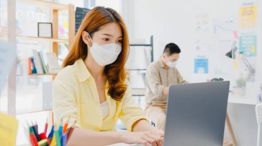 Asyalı kadınlar, dizüstü bilgisayar kullanırken ve ofiste akrilik bölünme ile ayrılırken, virüsün önlenmesi için yeni normal durumlarda sosyal mesafeler için yüz maskesi takarlar. Corona virüsünden sonra hayat ve iş.