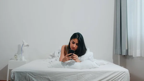 朝起きてからベッドに横になりながら Snsで幸せな笑顔を確認しながらスマホを使っている若いアジア人女性 自宅の寝室でリラックスした美しい魅力的なヒスパニック女性 — ストック写真