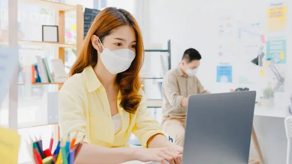 Asia female wear face mask for social distancing in new normal situation for virus prevention while using laptop and separated by acrylic partition stand in office. Life and work after corona virus.