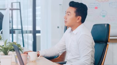 Başarılı yönetici Asyalı genç işadamı masaüstü bilgisayarı kullanarak gündelik giysiler giyer. Modern ofis işyerinde çalışma sırasında kaybolan ilham verici problem çözümü fikirleri düşünür..