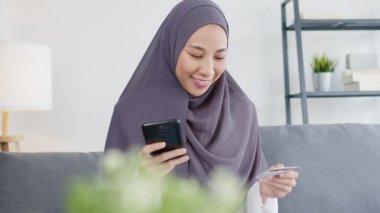 Asyalı Müslüman kadın akıllı telefon, kredi kartı satın alma ve evdeki oturma odasında e-ticaret interneti kullanıyor. Evde kal, online alışveriş, kişisel izolasyon, sosyal uzaklık, koronavirüs için karantina.