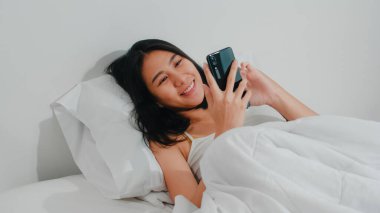 Genç Asyalı kadın akıllı telefon kullanıyor sosyal medyayı kontrol ediyor mutlu bir şekilde gülümsüyor sabah uyandıktan sonra yatakta yatıyor güzel, çekici İspanyol kadın gülümsüyor yatak odasında dinleniyor..