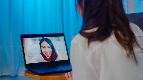 Asia student exchange female using laptop video call talking with friends while working from home at living room at night. Self-isolation, social distancing, quarantine for coronavirus in next normal.