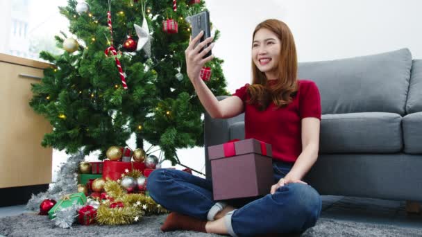 スマートフォンのビデオ通話を使用している若いアジアの女性は 自宅のリビングルームで装飾が施されたクリスマスツリー Masプレゼントボックスとカップルと話しています クリスマスと年末年始の祭り — ストック動画