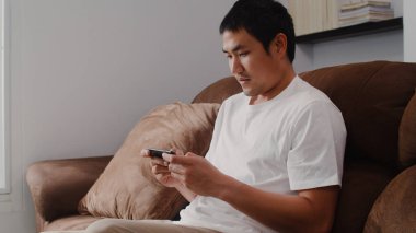 Genç Asyalı adam cep telefonu kullanıyor oturma odasında televizyon oyunu oynuyor, erkek kendini mutlu hissediyor evdeki kanepede uzanmış dinlenme zamanını kullanıyor. Erkekler ev kavramında oyun oynar rahatlar..
