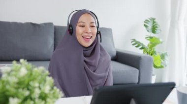 Asyalı Müslüman kadın, evdeki oturma odasında çalışırken konferans görüntülü konuşmasında iş arkadaşlarına satıştan bahsederek kulaklık takıyor. Corona virüsü için sosyal uzaklık, karantina.