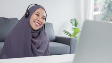Asya 'lı müslüman bayan, evdeki oturma odasında çalışırken iş arkadaşlarıyla konferans görüntülerinde satış raporları hakkında konuşarak kulaklık takıyor. Corona virüsü için sosyal uzaklık, karantina.