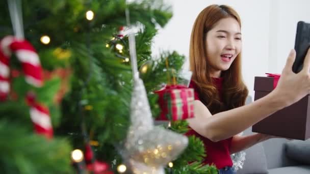 스마트 사용하는 아시아 여성이 거실에 장식이 크리스마스 트리인 상자를 부부와 — 비디오
