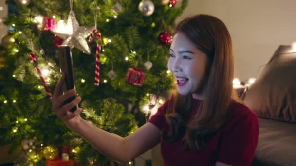 Masプレゼントボックス 自宅のリビングルームで装飾が施されたクリスマスツリーとカップルと話すスマートフォンのビデオ通話を使用して若いアジアの女性 クリスマスの夜と新年の休日の祭り — ストック動画