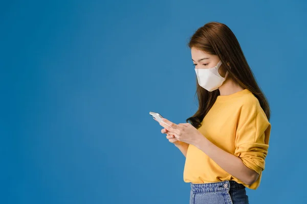 Young Asia girl wearing medical face mask using mobile phone with dressed in casual clothing isolated on blue background. Self-isolation, social distancing, quarantine for corona virus prevention.