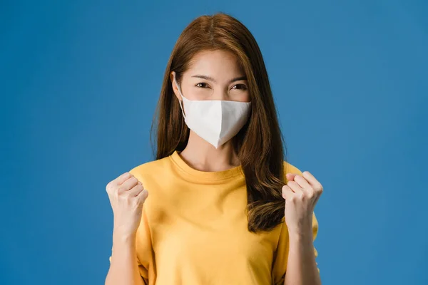 Young Asia girl wearing medical face mask showing peace sign, encourage with dressed in casual cloth and looking at camera isolated on blue background. Social distancing, quarantine for corona virus.