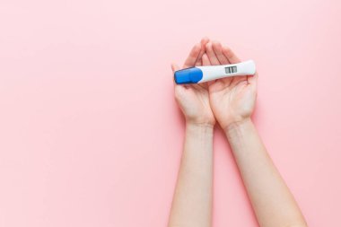 closeup of female hands holding positive pregnancy test with two stripes on pink background