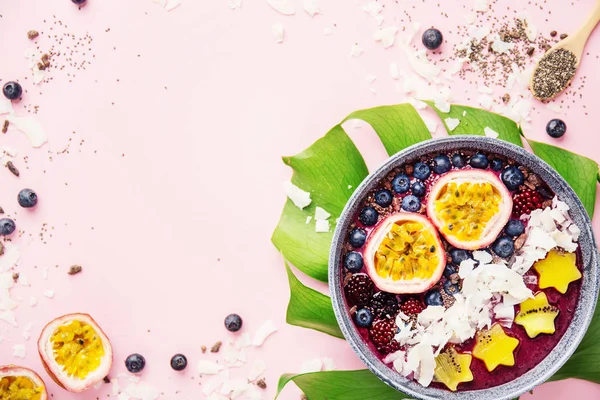 Tasty appetizing smoothie acai bowl made wild berries, decorated with cut passion fruit, coconut flakes, and cacao nibs on pink background