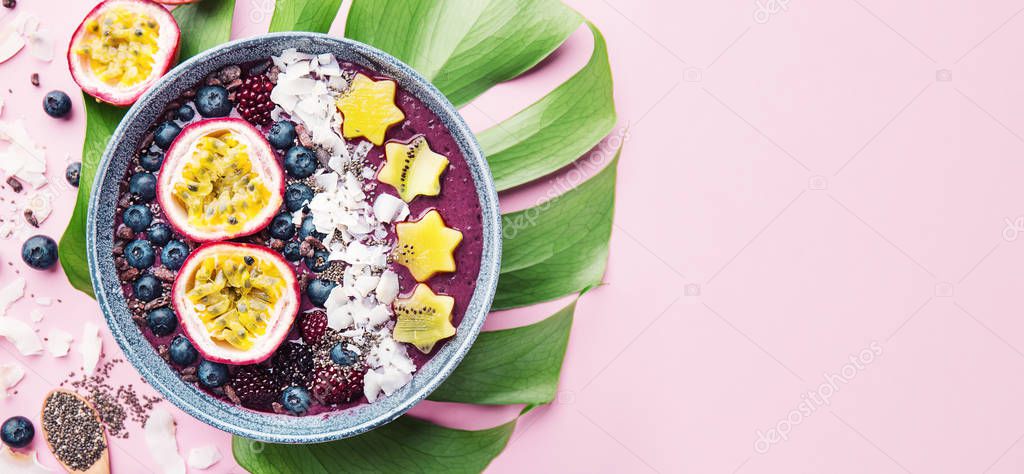 Tasty appetizing smoothie acai bowl made wild berries, decorated with cut passion fruit, coconut flakes, and cacao nibs on pink background