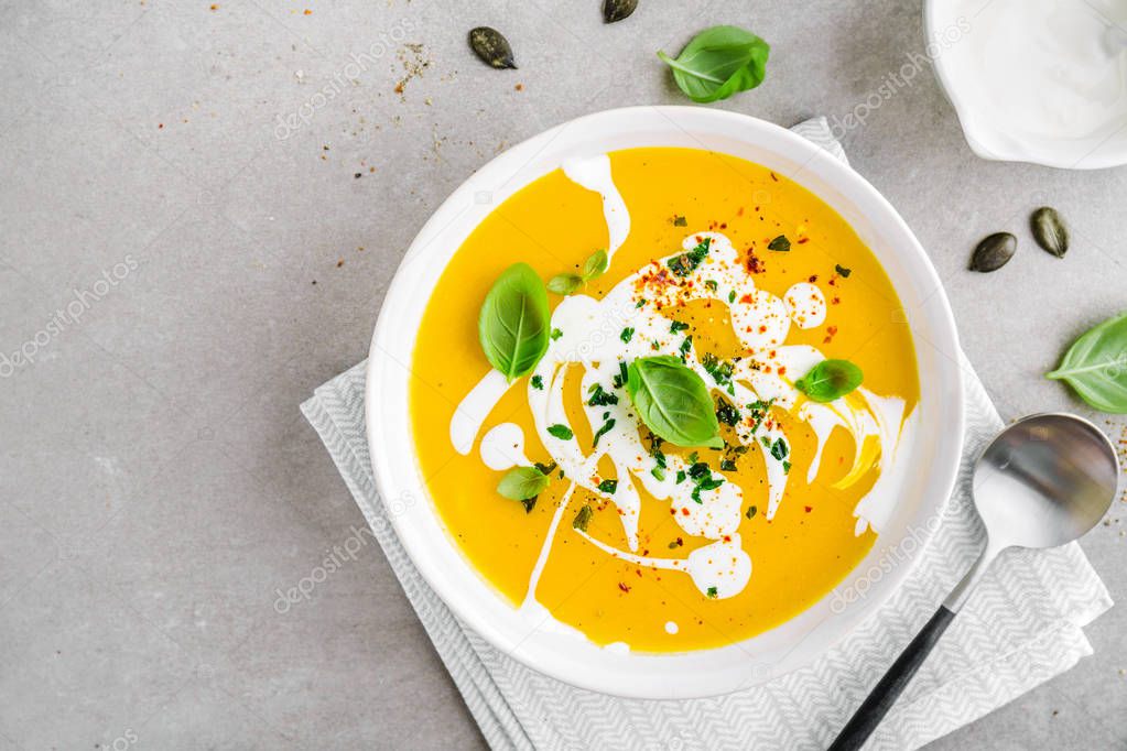 Tasty appetizing pumpkin vegetable creamy soup decorated with basil, cream and spices served in white bowl on grey table