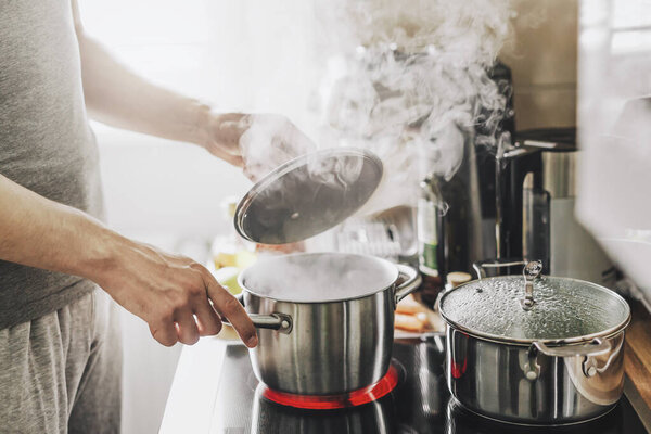 Young man cooking fresh food at home and opening lid of steaming pot. 