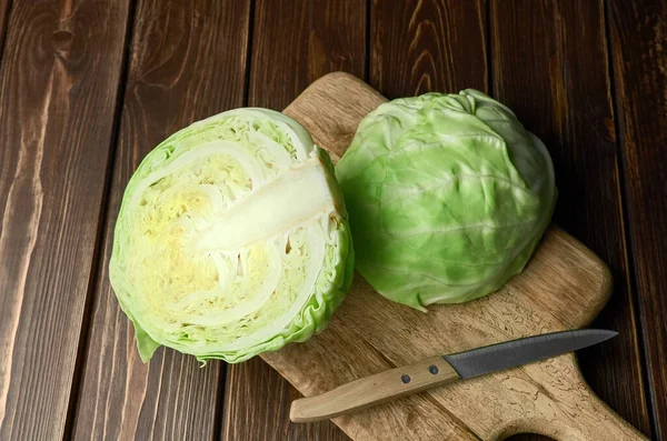 Halved fresh cabbage head with knife on cutting board over wooden table background. — Stock Photo, Image
