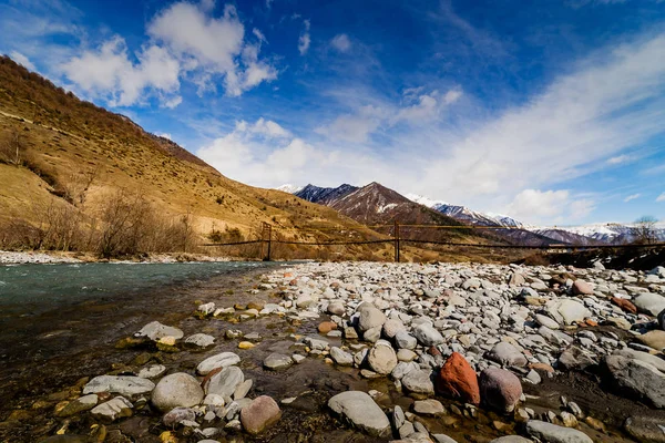 Dağ nehir kaya ve kayalık ve karla kaplı dağlar arka plan üzerinde asma köprü ile hızlı akışı ile. Gürcü askeri yola manzara.