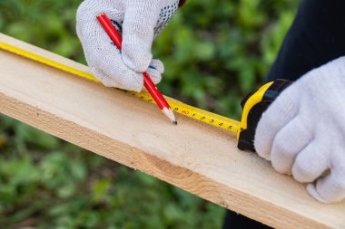 İş eldivenli bir marangoz, tahta üzerinde iz bırakmak için kalem ve bant mezurası kullanır. İnşaat endüstrisi, marangozluk atölyesi.