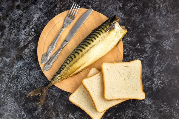 One smoked mackerel without head with fork knife cutting board bread on black background