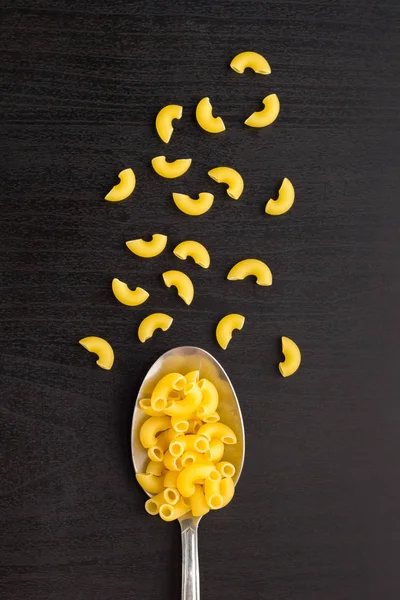 Cuernos de pasta cruda italiana seca en una cuchara metálica y una mesa negra — Foto de Stock