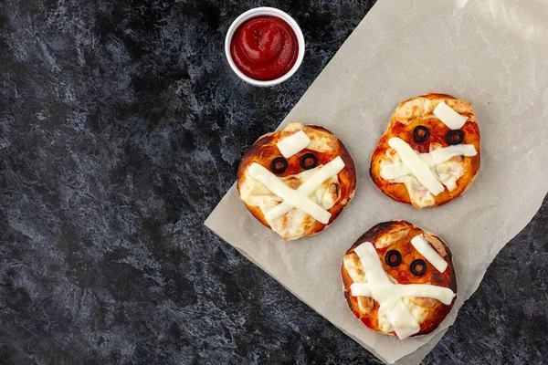Peynirli, zeytinli ve ketçaplı çocuklar için mumya olarak mini pizza. Karanlık arka plandaki çocuklar için komik çılgın Cadılar Bayramı yemekleri