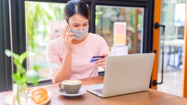 Asyalı kadın yüz maskesi takıyor ve online alışveriş yapıyor. Sosyal uzaklık ve yeni normal yaşam tarzı.