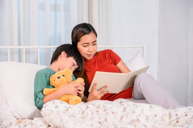 Asyalı anne oğluyla birlikte evde çalışıyor. Anne ve çocuk yatakta uyumadan önce masal okuyorlar. Kadın yaşam tarzı ve aile aktiviteleri.