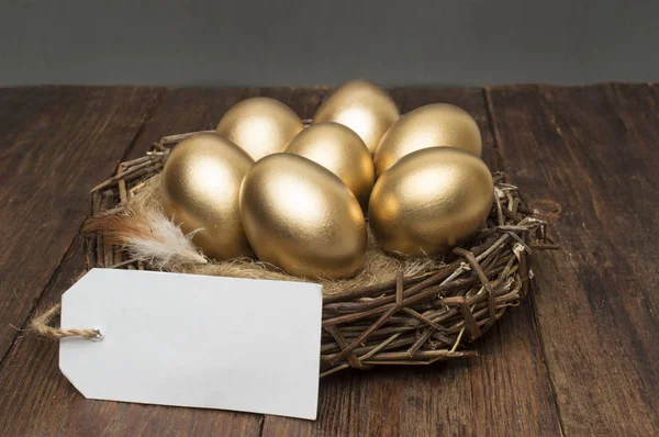 Nido con huevos dorados con una etiqueta y lugar para texto sobre un fondo de madera. El concepto de jubilación exitosa —  Fotos de Stock