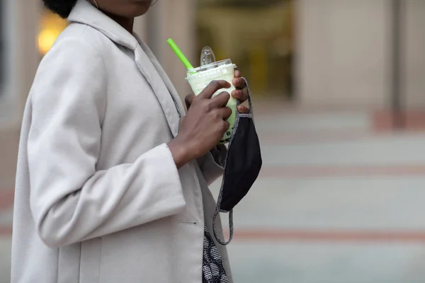 A fabric face mask and a drink in the hand of a black person. Face masks are recommended to wear to prevent the spread of coronavirus pandemic.