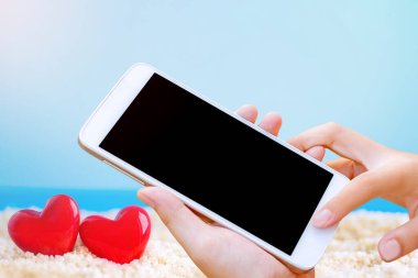 Kadın el ele tutuşup ekrandaki akıllı telefona ya da cep telefonuna plaj arka planındaki kırmızı kalbe dokunuyor..