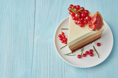 Açık mavi ahşap bir masada kırmızı kuş üzümü, incir, tarçın ve biberiye ile süslenmiş ev yapımı kek bir parça.