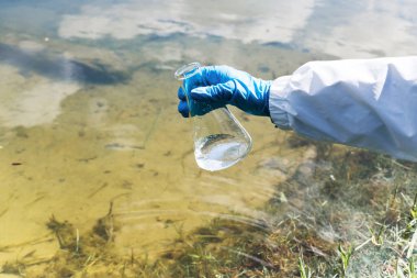 Çit, su örneğinde enfeksiyon olup olmadığına bak. Elinde bir matara ve gölün, nehrin ve denizin arka planında test tüpü olan eldivenli bir el. Ekoloji ve çevre kirliliği
