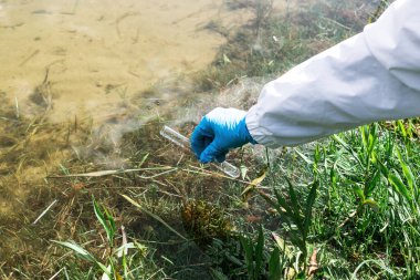 Çit, su örneğinde enfeksiyon olup olmadığına bak. Elinde bir matara ve gölün, nehrin ve denizin arka planında test tüpü olan eldivenli bir el. Ekoloji ve çevre kirliliği