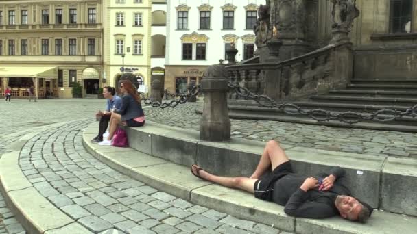 OLOMOUC, REPÚBLICA CHECA, 1 DE SEPTIEMBRE DE 2018: Auténtico hombre emocionado dormido en la columna de la plaga del patrimonio de la UNESCO, se sienta con los turistas y camina por la plaza, la vida en la ciudad — Vídeos de Stock