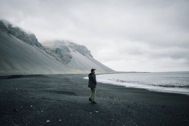 Lonely man walks on grey black volcanic sand beach clipart