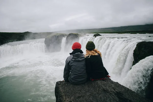 Пара сидит на краю скалы у огромного водопада — стоковое фото